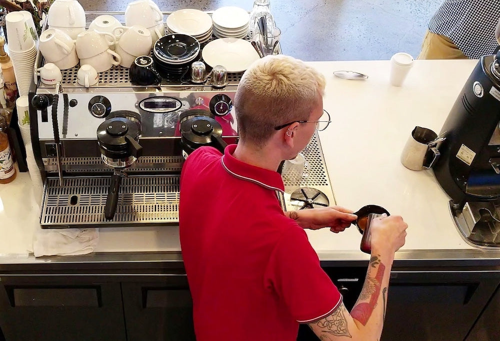 Barista in a red shirt making a latte art espresso drink while standing in front of a La Marzocco Strada espresso machine.