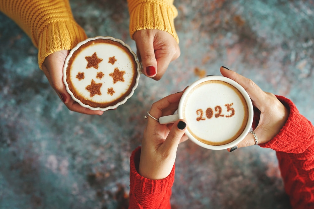 Pair of female hands holding two coffees. One with 2025 on the coffee and the other with stars on the coffee.