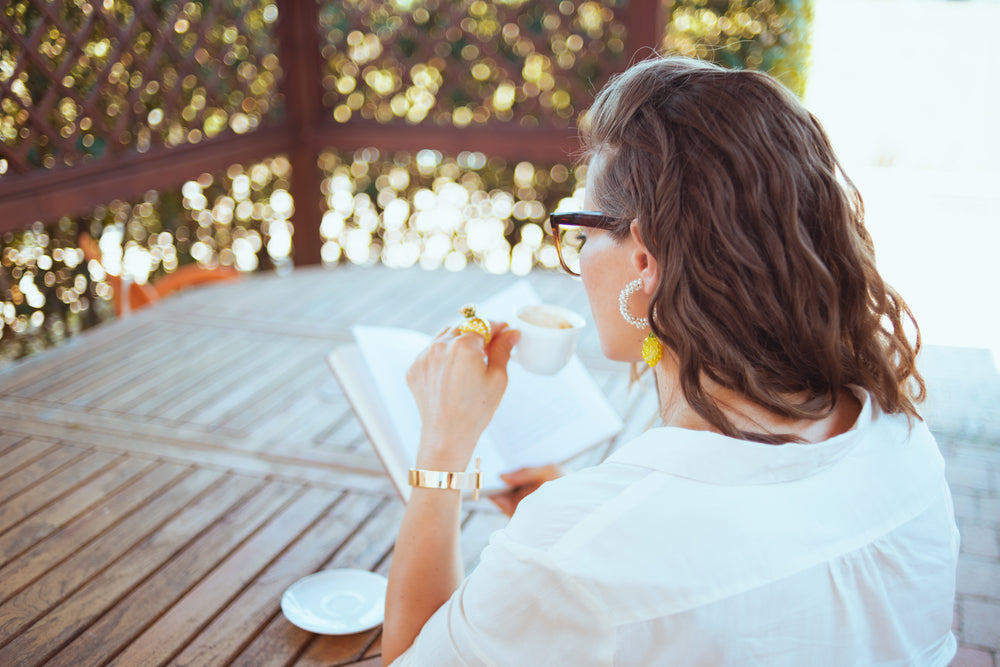 Seen from behind woman in white shirt with eyeglasses sitting at the table drinking coffee and reading book in the terrace.