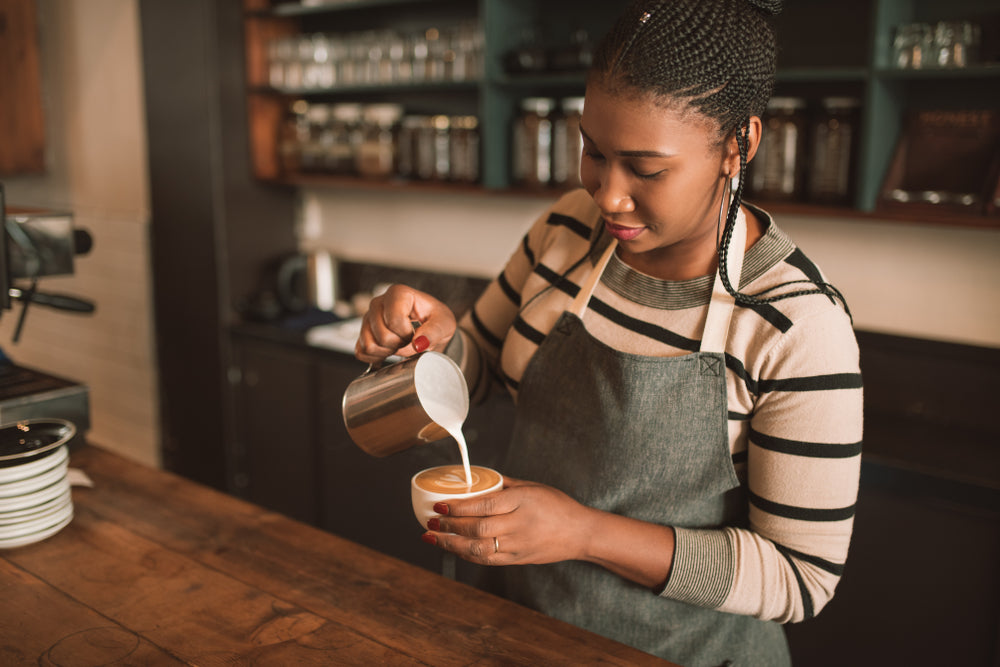 Smiling young female barista standing behind the counter of her trendy cafe pouring frothed milk into a cup of cappuccino.