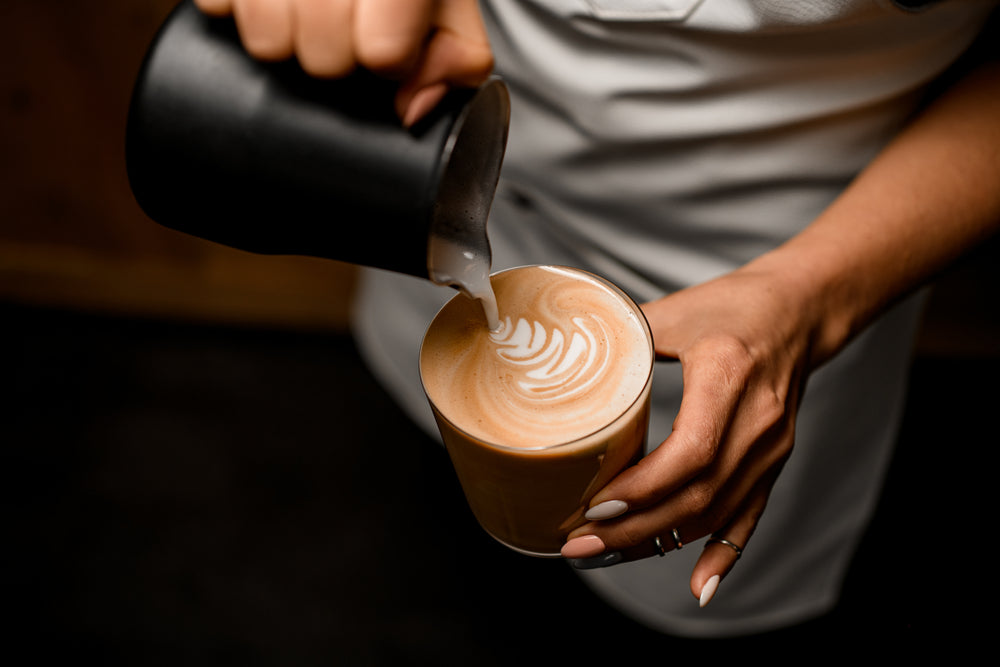 Woman pours milk from jug into coffee in a glass to make latte art.
