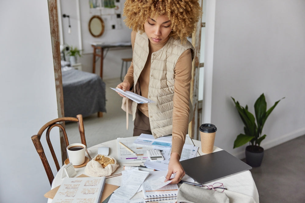 Cropped shot of young woman checks bills calculates expenses stands near table against cozy home interior.