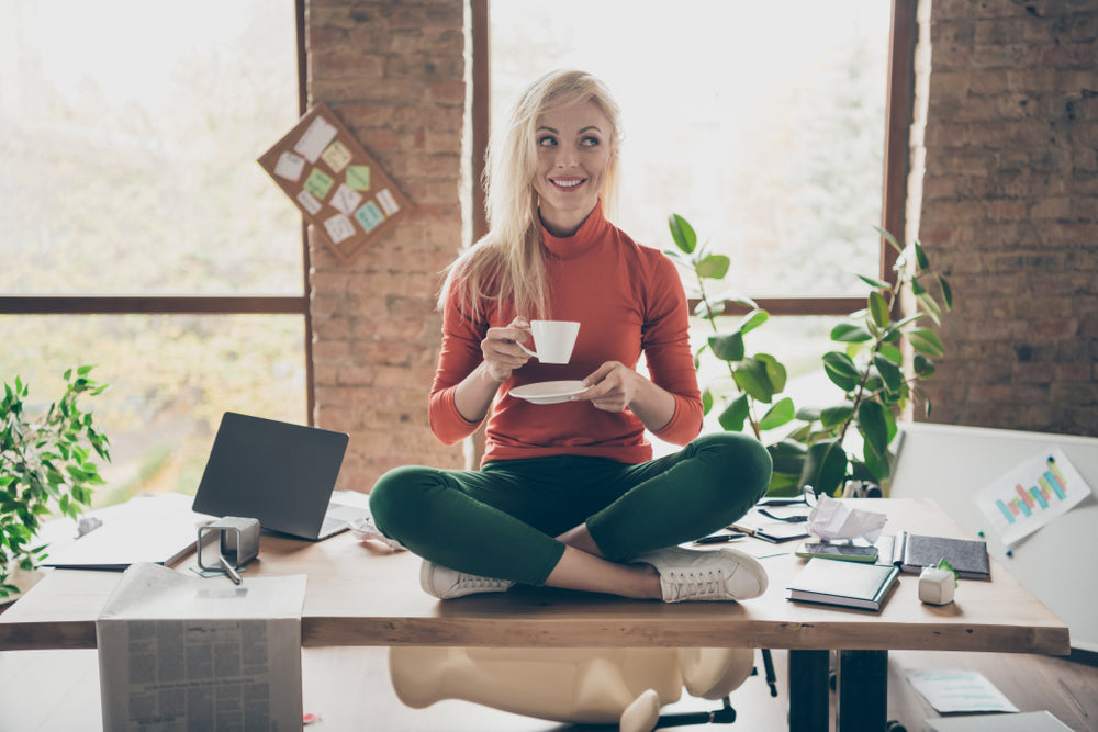 Full body photo of happy positive woman company owner sit on table crossed legs rest relax hold coffee cup drink cappuccino wear red turtleneck green pants trousers in messy office loft.