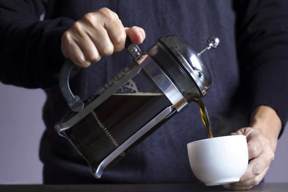 Man in blue long sleeve shirt pours coffee from french press into a white coffee cup.