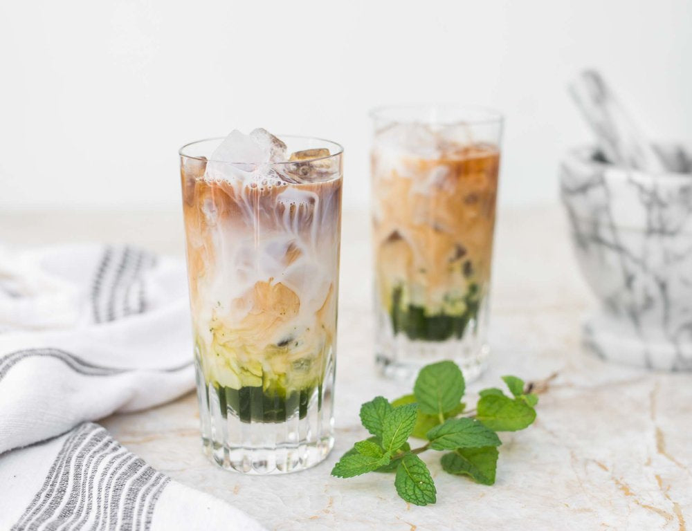 Two glasses of Mint Iced Latte with dishtowels in the background and mint leaves in the foreground.