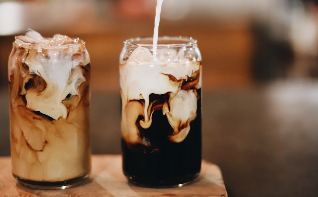 Two glasses of cold brew on a wooden table with milk pouring into one glass.