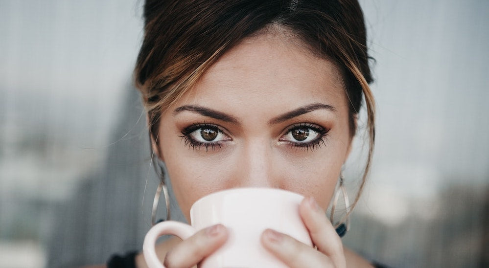 Woman with brown eyes and hair stares while holding white cup with both hands as she drinks.