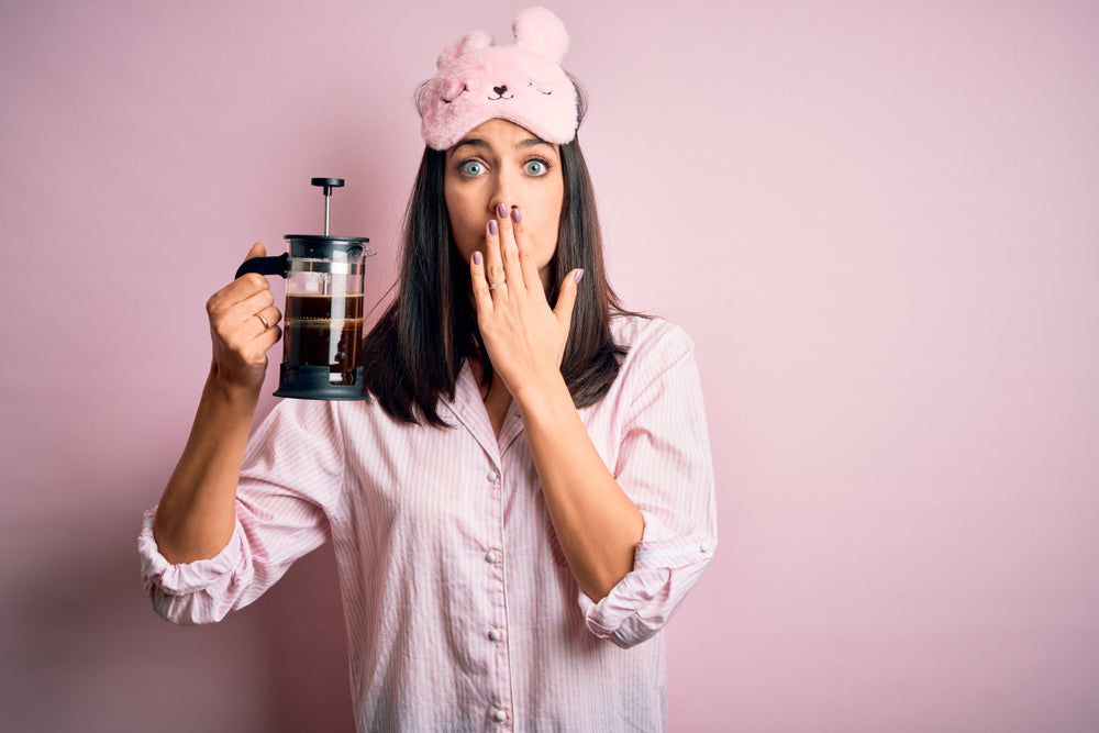 Young brunette woman with blue eyes wearing pajama making coffee with french press covers her mouth with hand shocked with shame for mistake with coffee preparation.