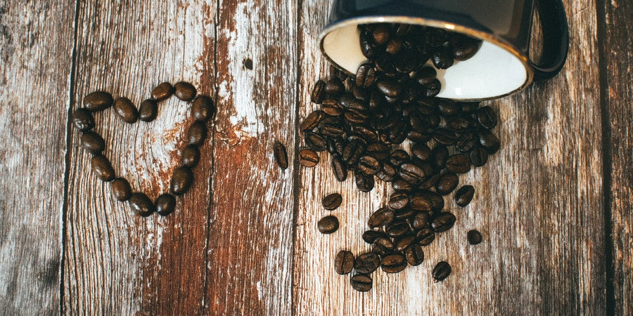 Roasted coffee beans shaped into a heart next to a brown coffee mug spilling roasted coffee beans across a wood table.